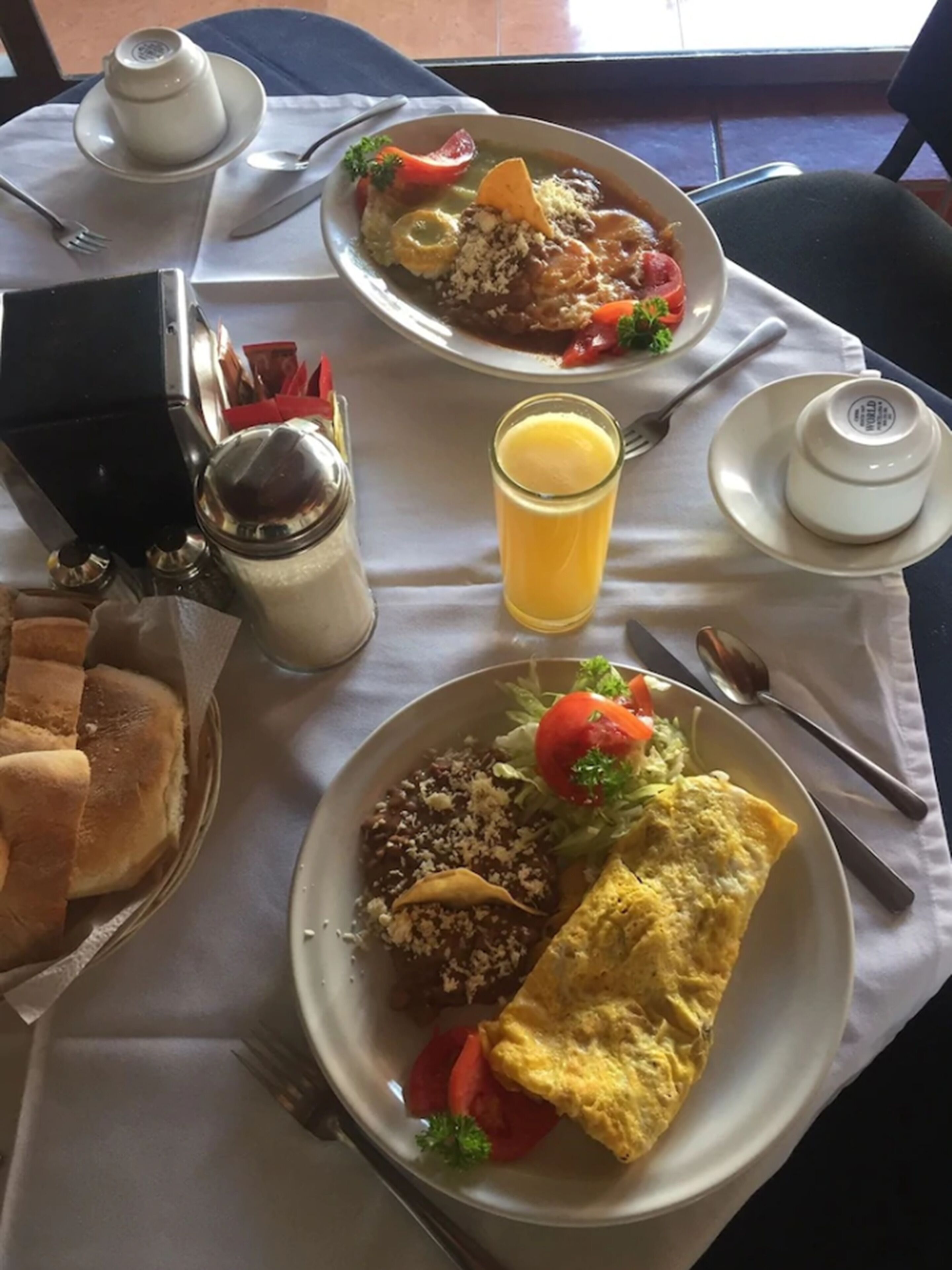 Uma mesa posta com dois pratos de comida, um copo de suco de laranja, saleiros e pimenteiros, uma cesta de pão e uma pequena tigela.
