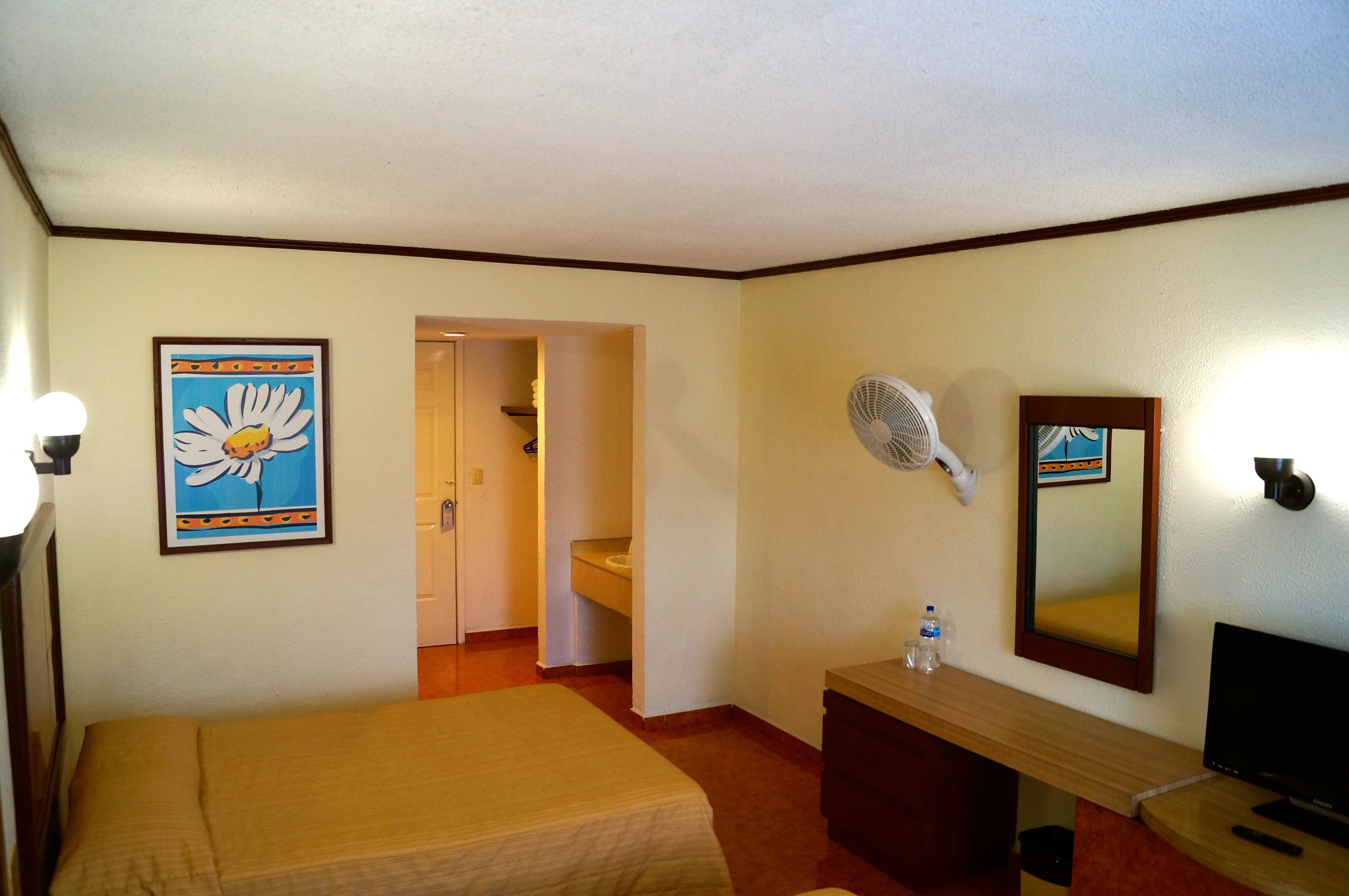 A hotel room with a bed, a desk with a mirror, a fan, and a framed picture on the wall.
