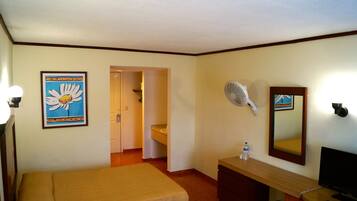 A hotel room with a bed, a desk with a mirror, a fan, and a framed picture on the wall.