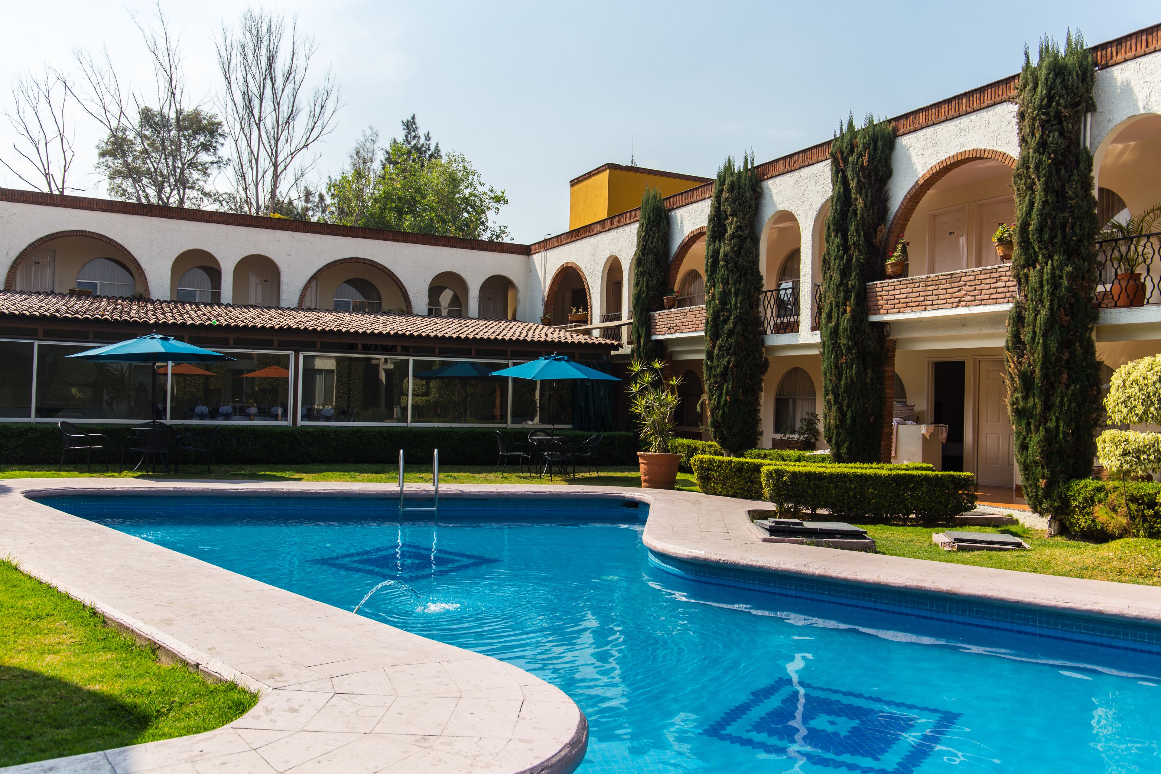 Uma piscina com deck ladrilhado, cercada por um edifício com janelas em arco e telhado ladrilhado.