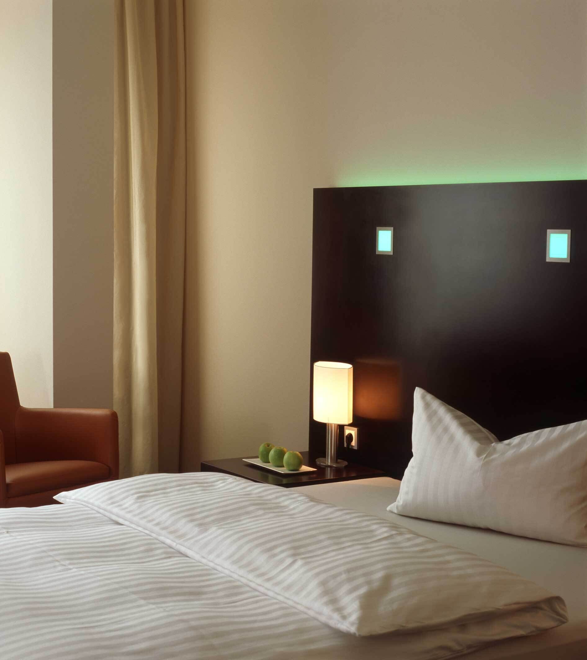 Une chambre d’hôtel avec un lit, une table de chevet avec une lampe, une chaise et une tête de lit sombre agrémentée de deux petites lampes.