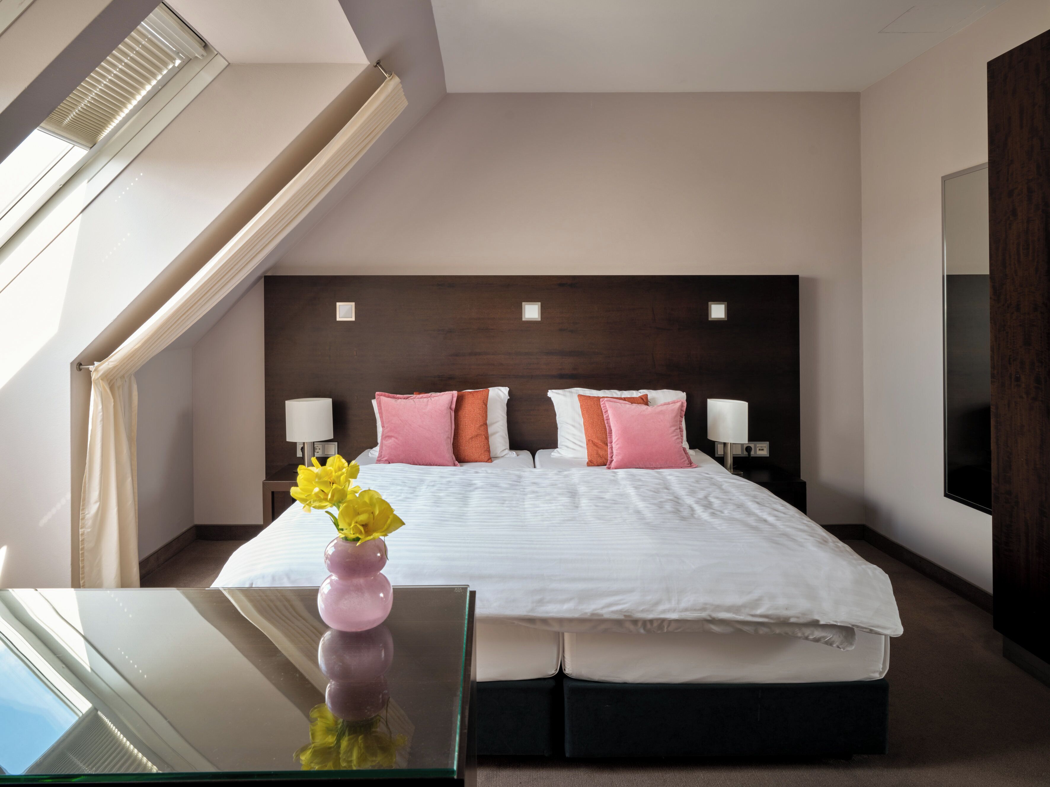 A modern hotel room with a large bed, bedside lamps, a glass table with flowers, and a view of the sky through a window.