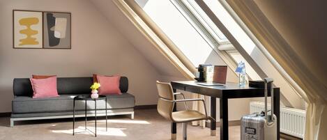 A modern loft bedroom with a skylight, a gray sofa, a small table, a chair, and a suitcase.