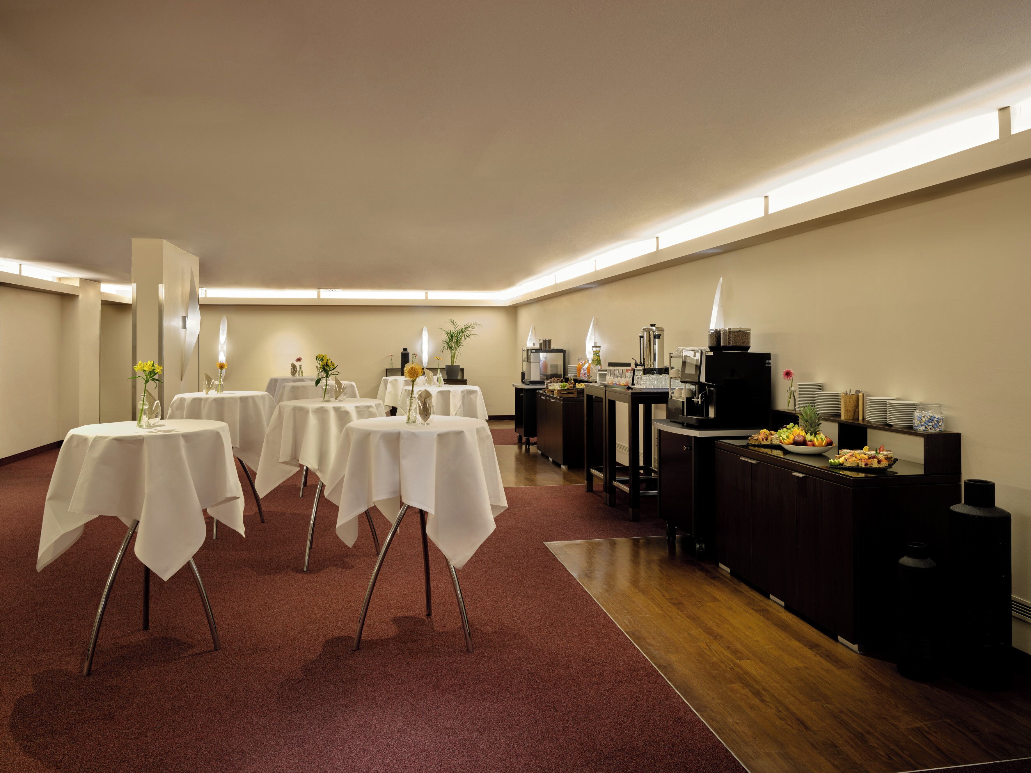 A conference room with multiple tables covered with white tablecloths, equipped with coffee machines and a buffet area with fruits and snacks.