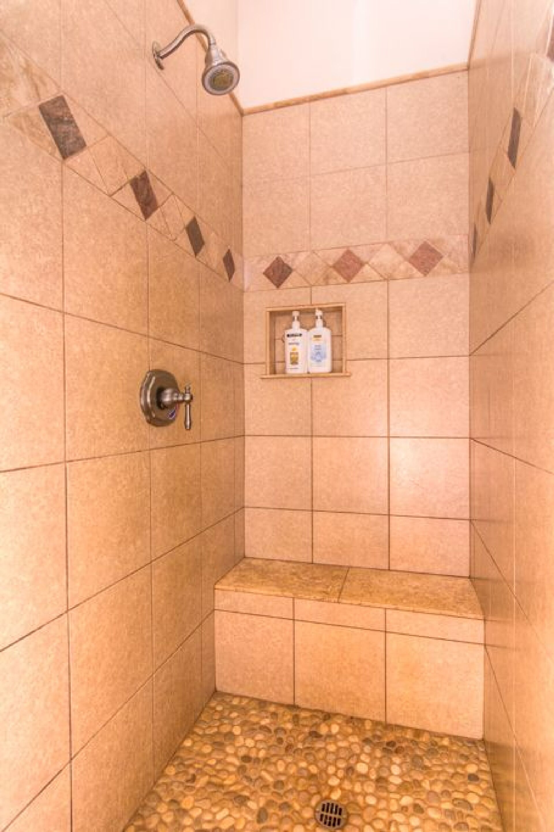 A shower with a tiled wall, a showerhead, a shelf with toiletries, and a pebble-patterned floor.