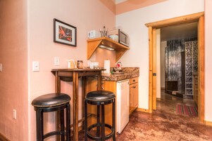 Cocina compacta con microondas, tostadora y cafetera sobre una estante de madera. Hay dos taburetes y un refrigerador.
