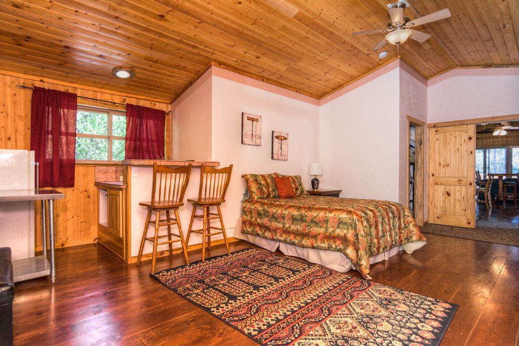 A cozy bedroom with a wooden ceiling, a bed with a patterned bedspread, two wooden chairs, a small table, and a window with red curtains.