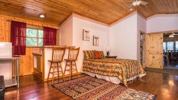 Un dormitorio acogedor con techo de madera, una cama con colcha estampada, dos sillas de madera, una mesita y una ventana con cortinas rojas.
