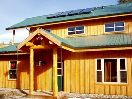 A two-story wooden building with a green roof, solar panels, and a covered porch with a swing set.