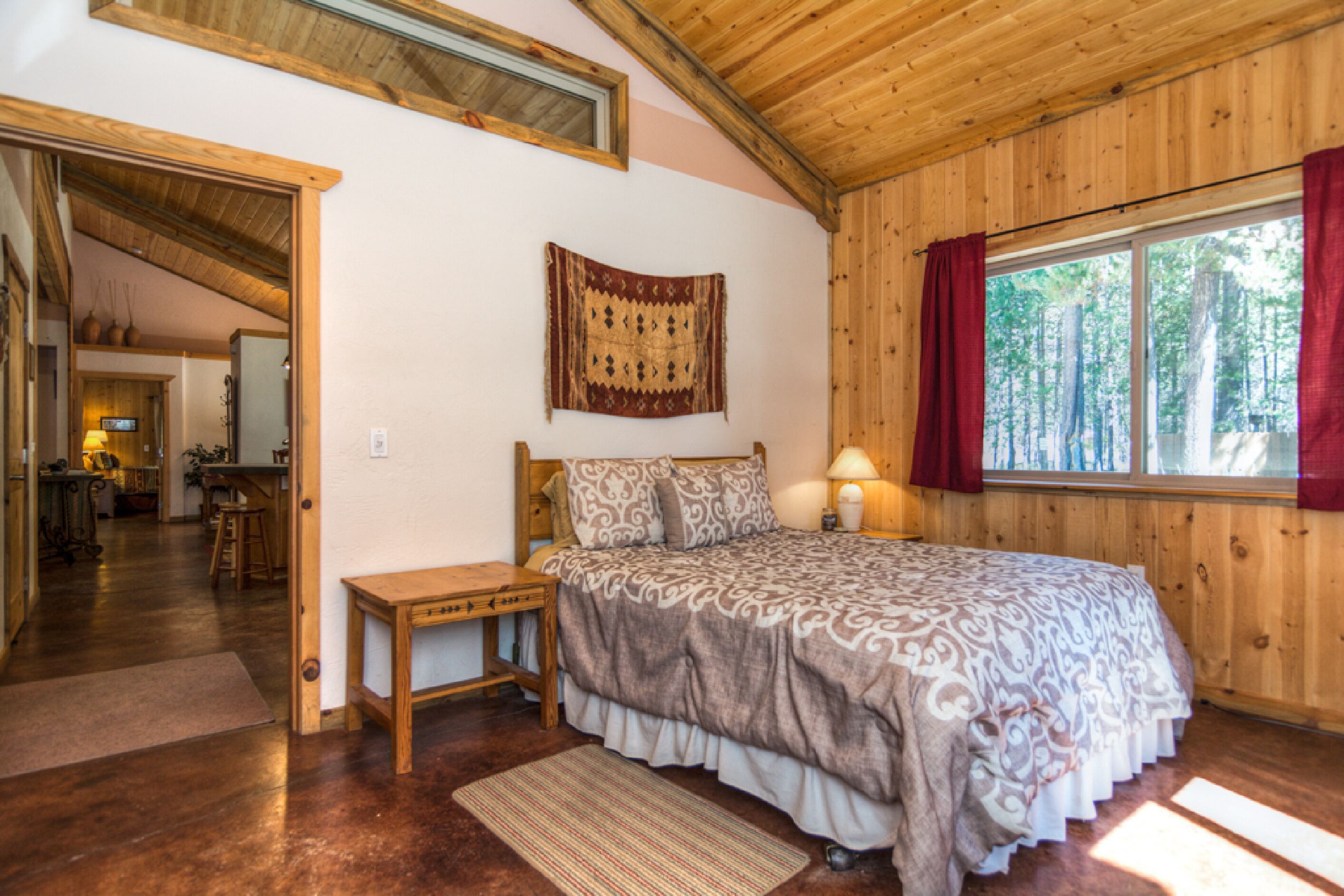 Una camera da letto con soffitto in legno, un letto grande con biancheria a motivi, un comodino, una finestra con tende e vista su alberi.
