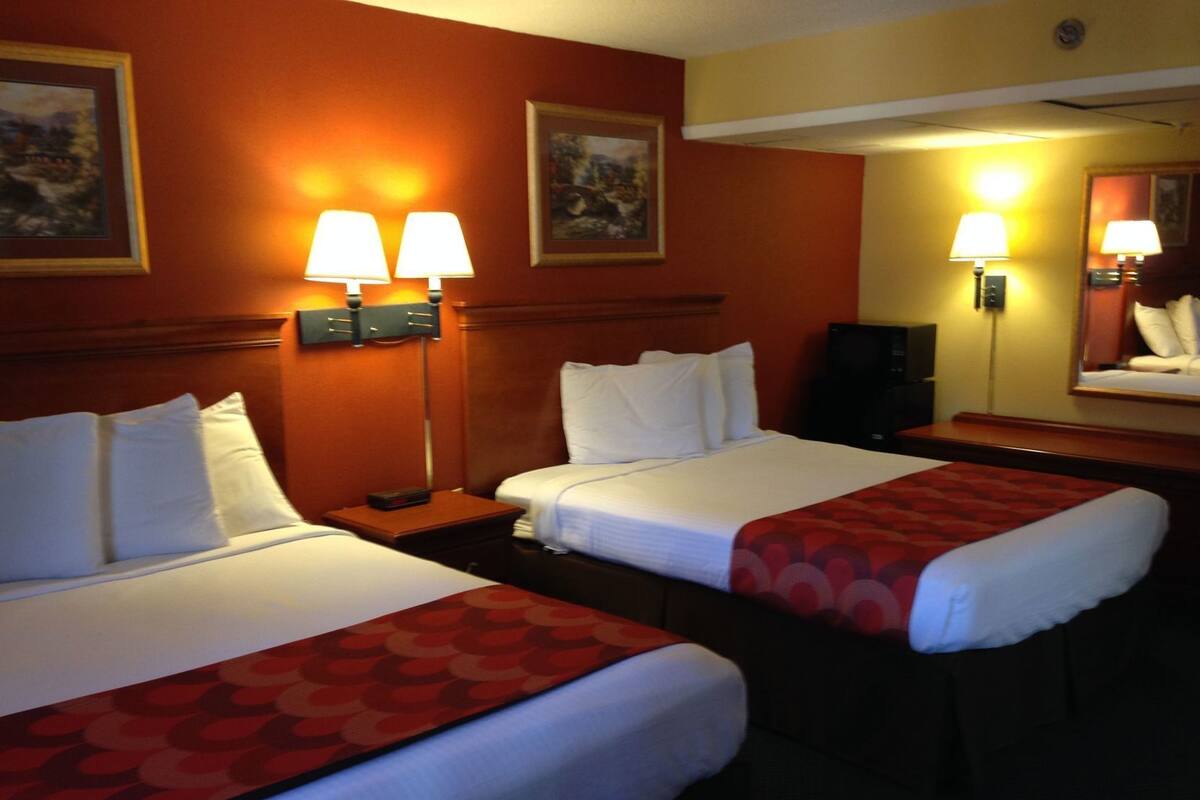 A hotel room with two beds, each with white linens and pillows, a red patterned bed runner, and two wall-mounted lamps.