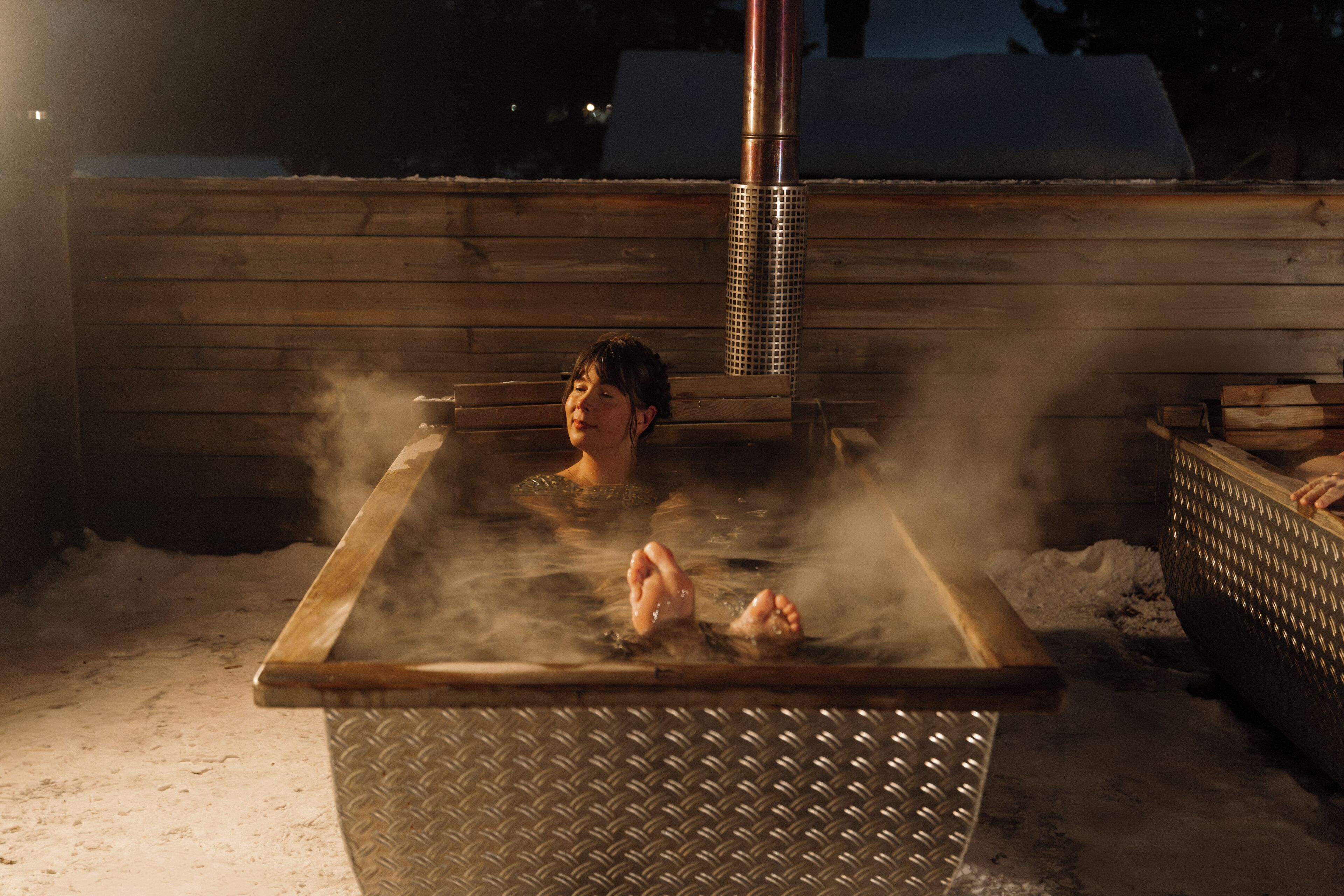 A person in a hot tub with steam rising, surrounded by a wooden deck and a metal structure.