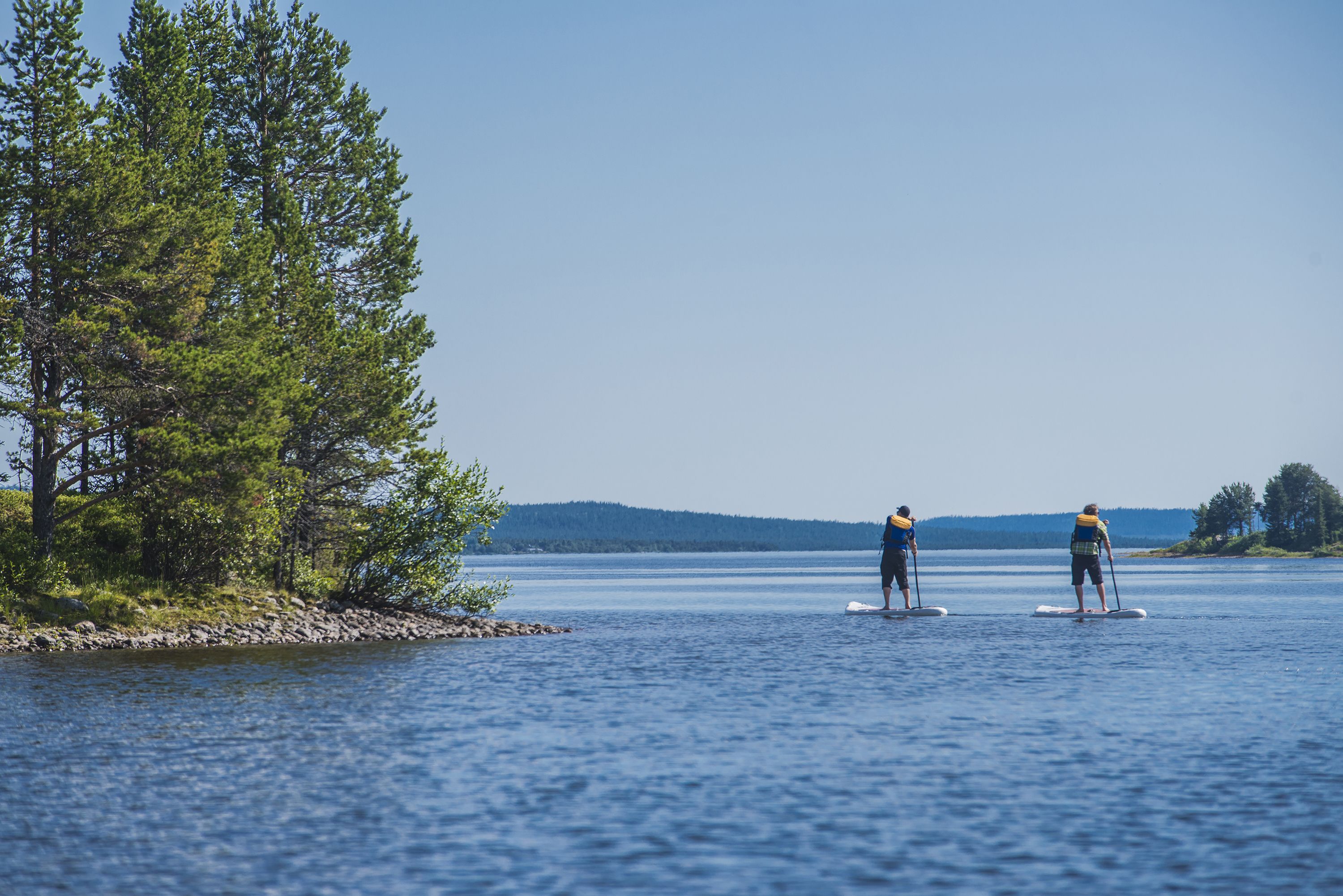 Zwei Personen stehen auf Paddleboards auf einem ruhigen See, im Hintergrund Bäume und Hügel.