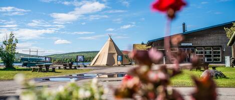 Edificio de recepción con un letrero, un tipi, mesas de picnic y un bote al fondo.