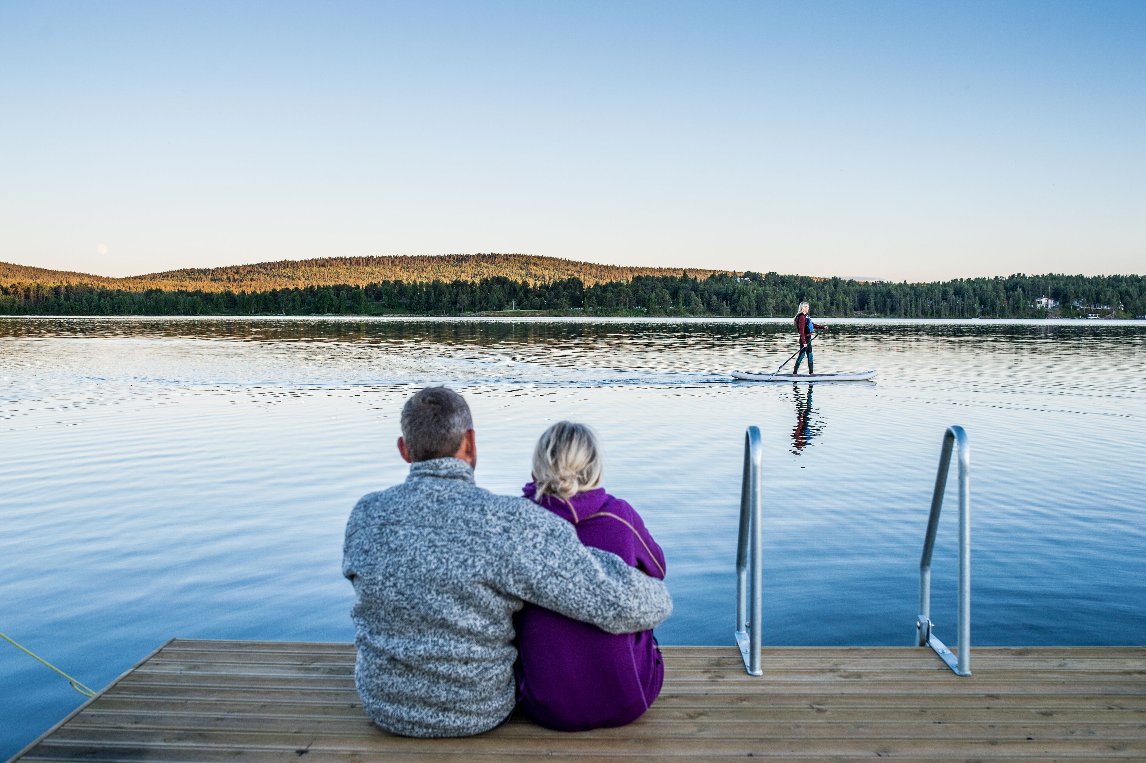 A couple sitting on a wooden dock overlooking a calm lake with a person paddleboarding in the distance.