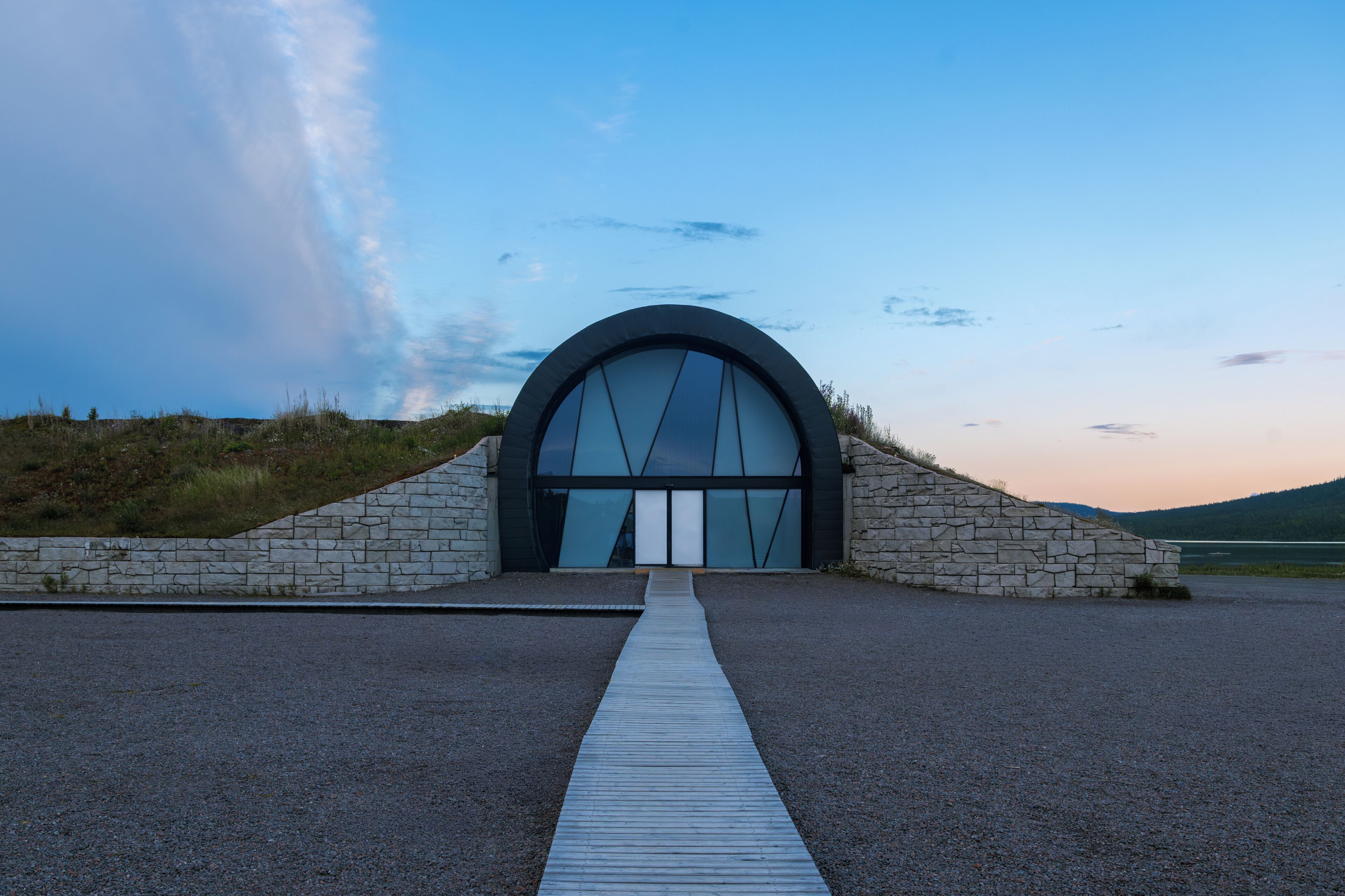 Un bâtiment moderne doté d’une grande entrée en arc de cercle, d’un chemin de pierre menant à l’entrée et, en arrière-plan, d’un ciel dégagé.