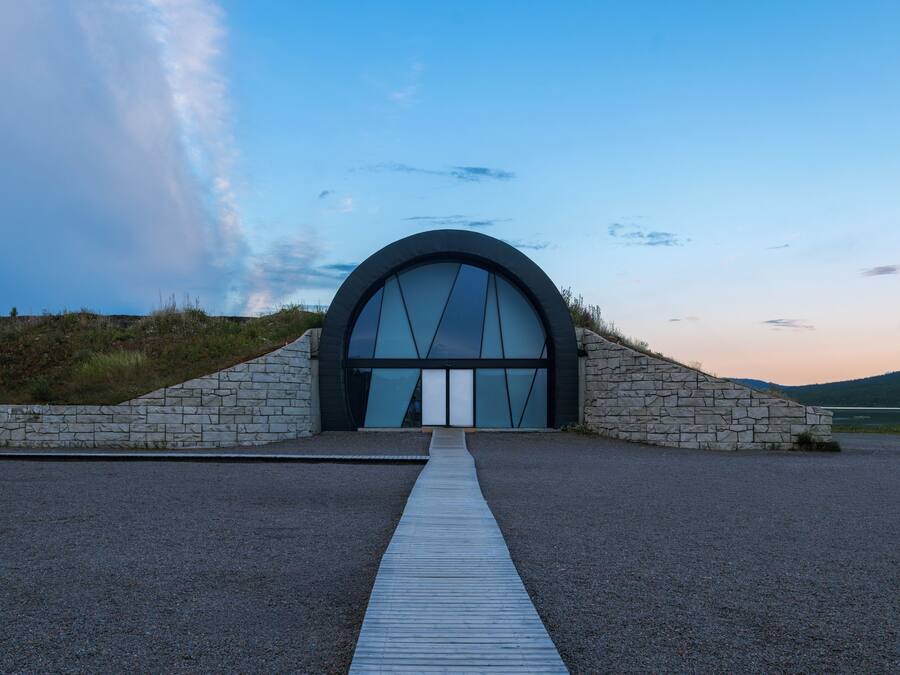 Een modern gebouw met een grote gebogen entree, een pad van stenen dat erheen leidt, en een heldere lucht op de achtergrond.