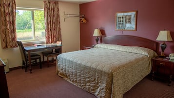 Una camera d'albergo con un letto grande, una scrivania, due lampade, una finestra con tende e un quadro appeso al muro.