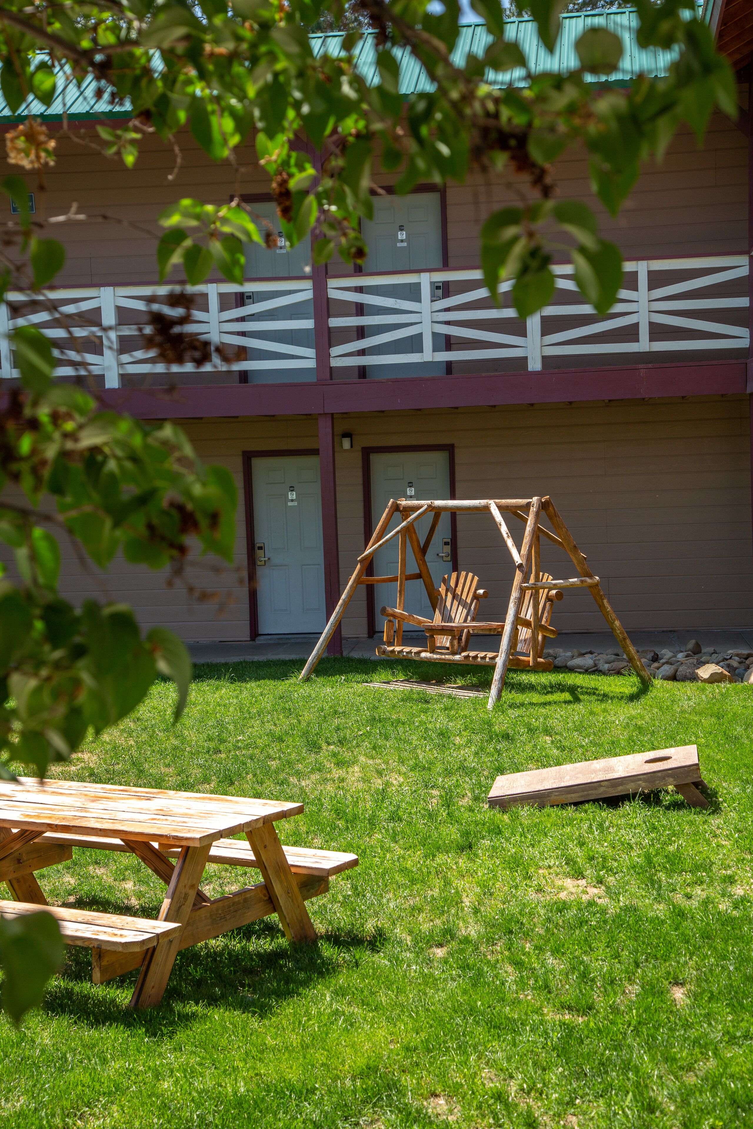 A wooden swing set and picnic table in a grassy area in front of a building with a white fence and a door.