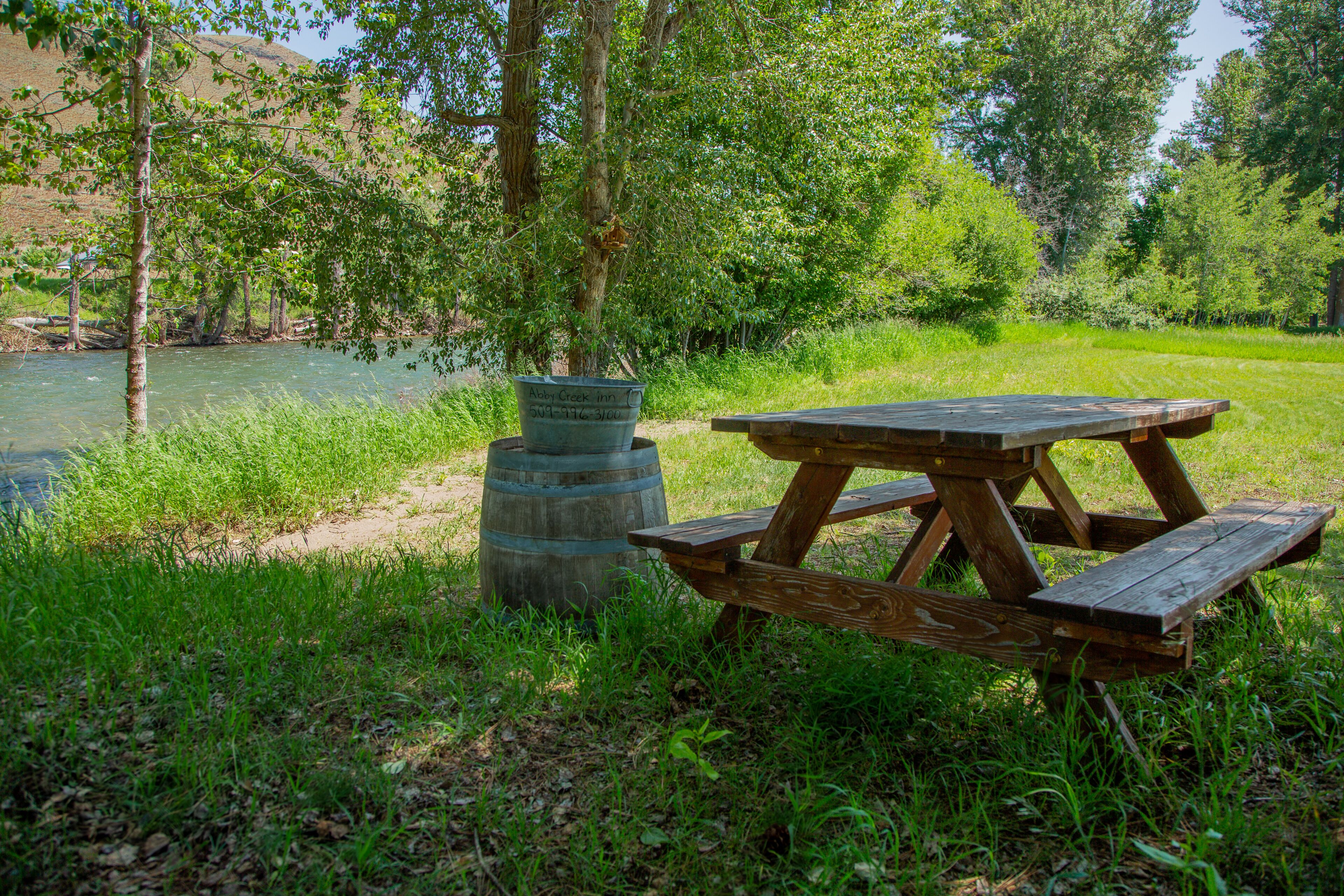 Una mesa de picnic y un barril con un cartel en un área verdejante cerca de un río.