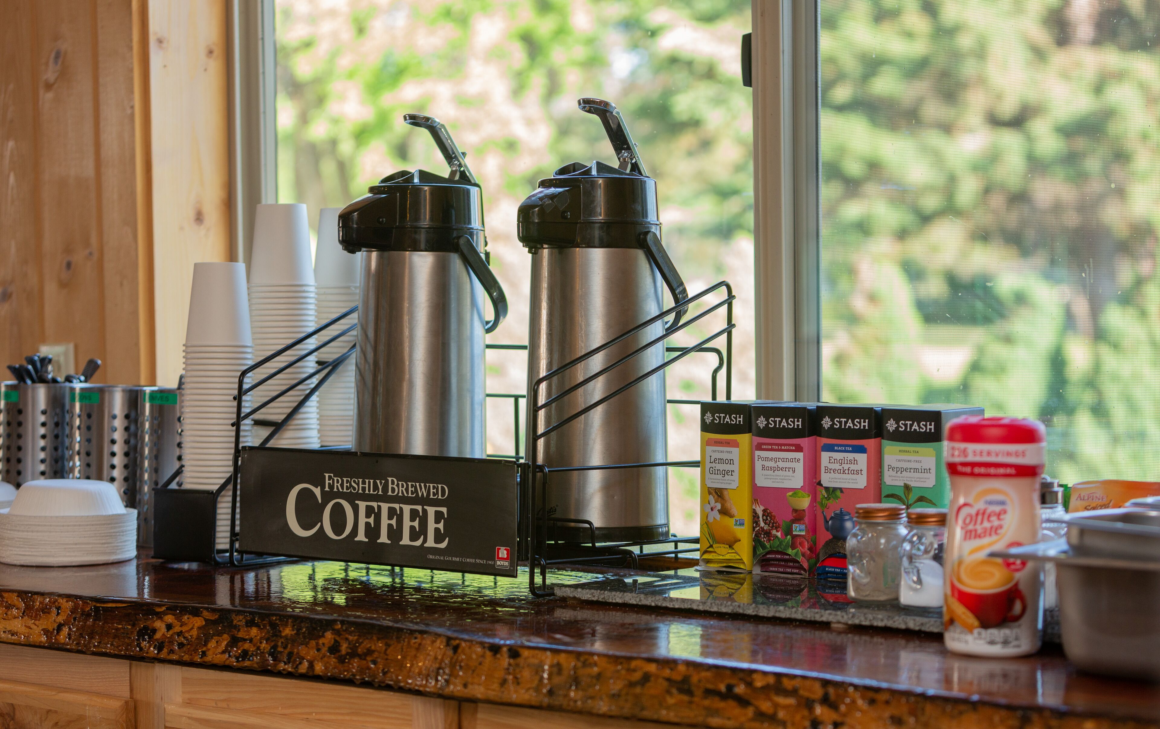 Una estación de café con dos máquinas de café de acero inoxidable, un letrero que dice "Café Recién Hecho" y diversos productos de café y té sobre el mostrador.