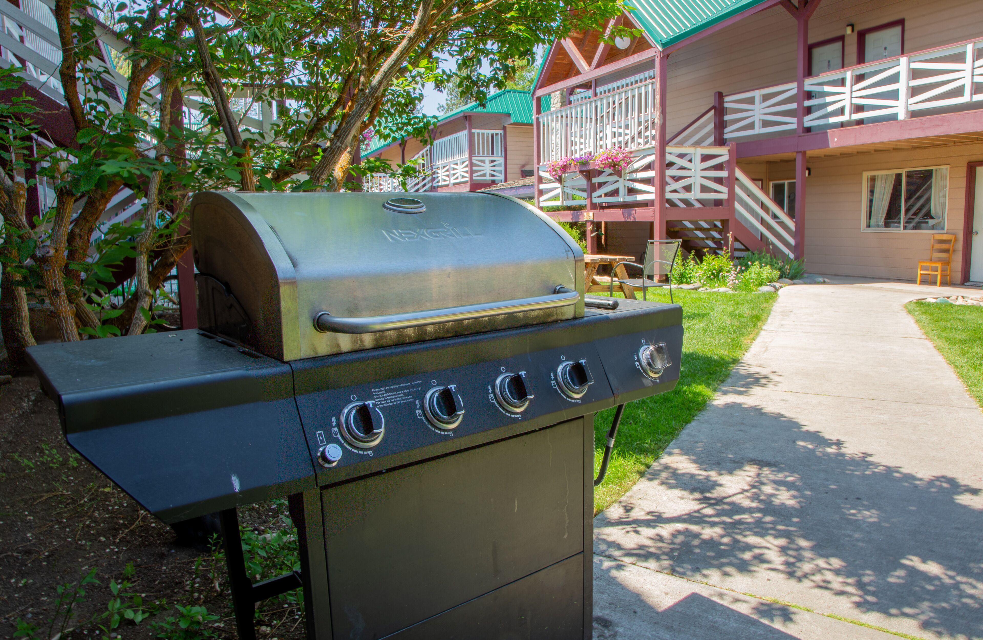 A barbecue grill with four burners, located in a courtyard with a building in the background.