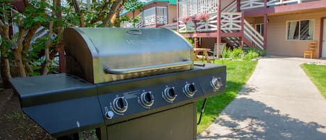 A barbecue grill with four burners, located in a courtyard with a building in the background.