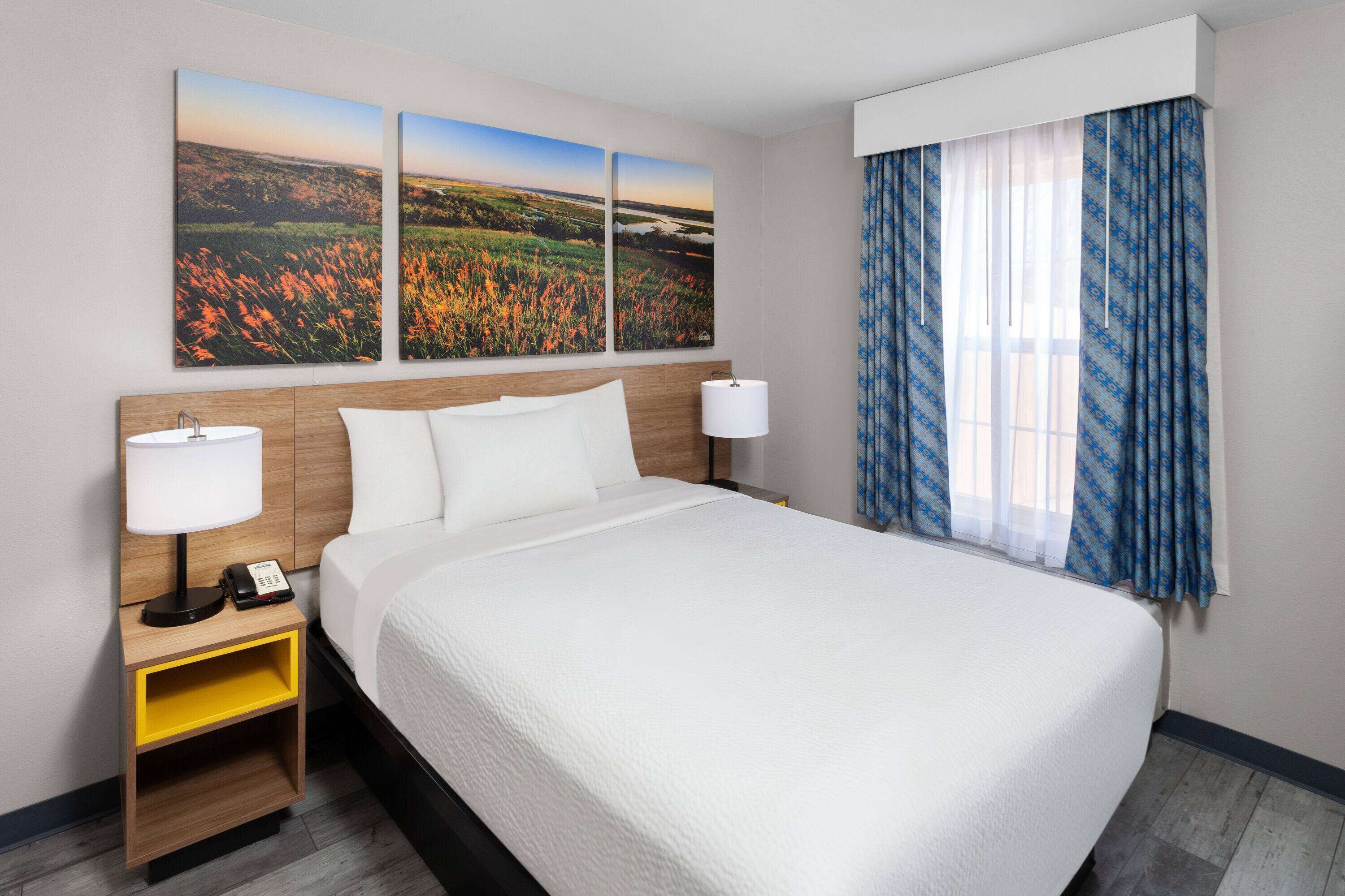 Una habitación de hotel con una cama grande, dos mesitas de noche con lámparas, una mesita con un teléfono y una ventana con cortinas azules.
