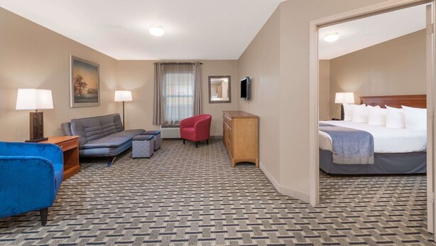 A hotel room with a patterned carpet, a blue armchair, a red chair, a sofa, a wooden dresser, a bed with white linens, and a flat-screen TV mounted on the wall. - Days Inn & Suites by Wyndham Sellersburg (Sellersburg)
