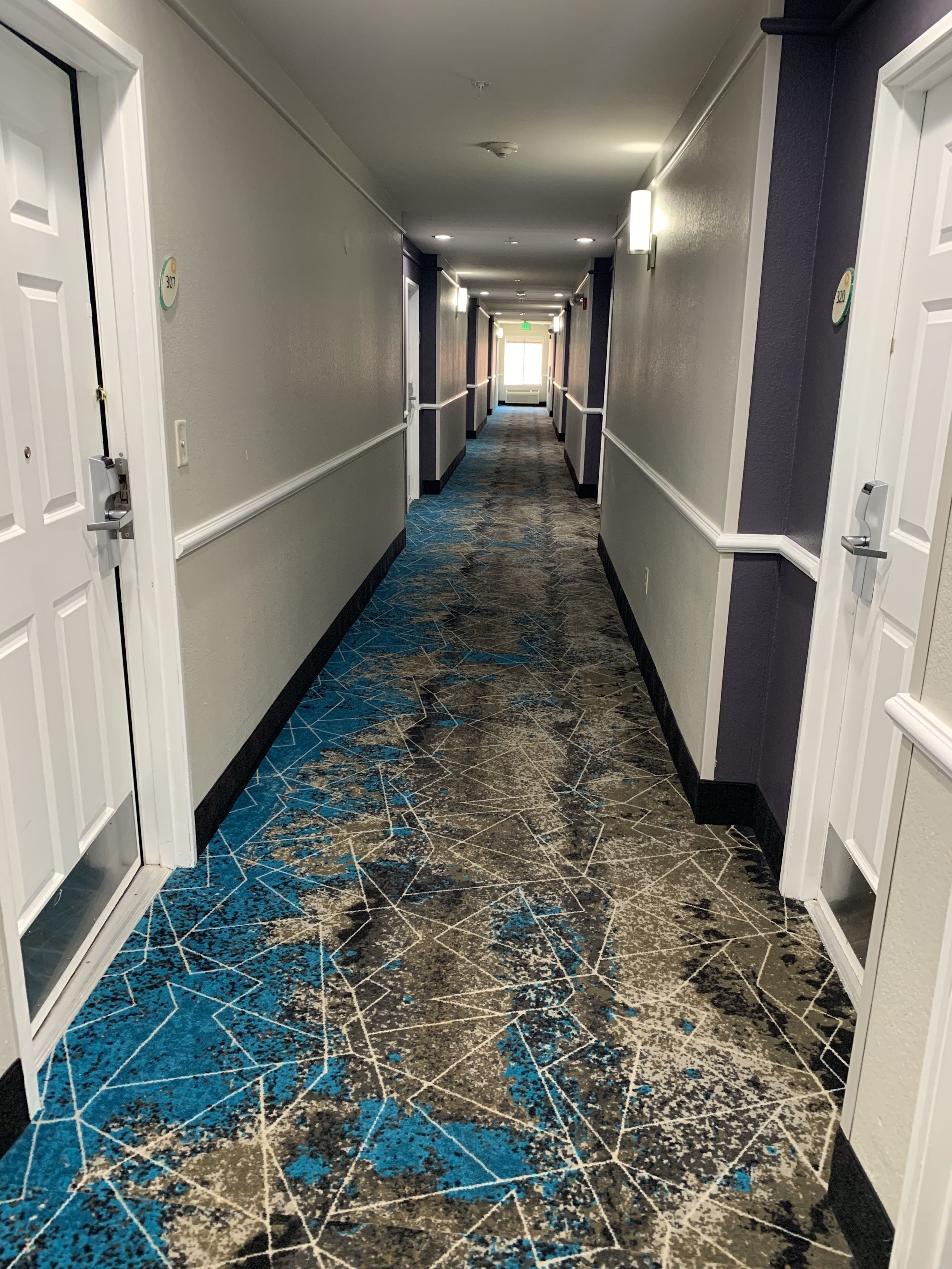 En hotellkorridor med ett mönstrat golv, vita dörrar och en lampa i slutet.