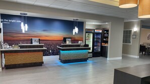 A reception area with two reception desks, a wall mural, a refrigerator stocked with beverages, and a seating area in the background. - La Quinta Inn & Suites by Wyndham Lubbock North (Lubbock)