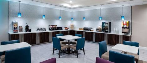 A modern restaurant with a central dining area featuring a white table surrounded by blue and purple chairs. There are hanging blue pendant lights above the tables, and a counter with various food items and a beverage dispenser.