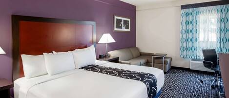 Quarto de hotel com uma cama grande, uma escrivaninha, uma cadeira, um abajur e cortina estampada.