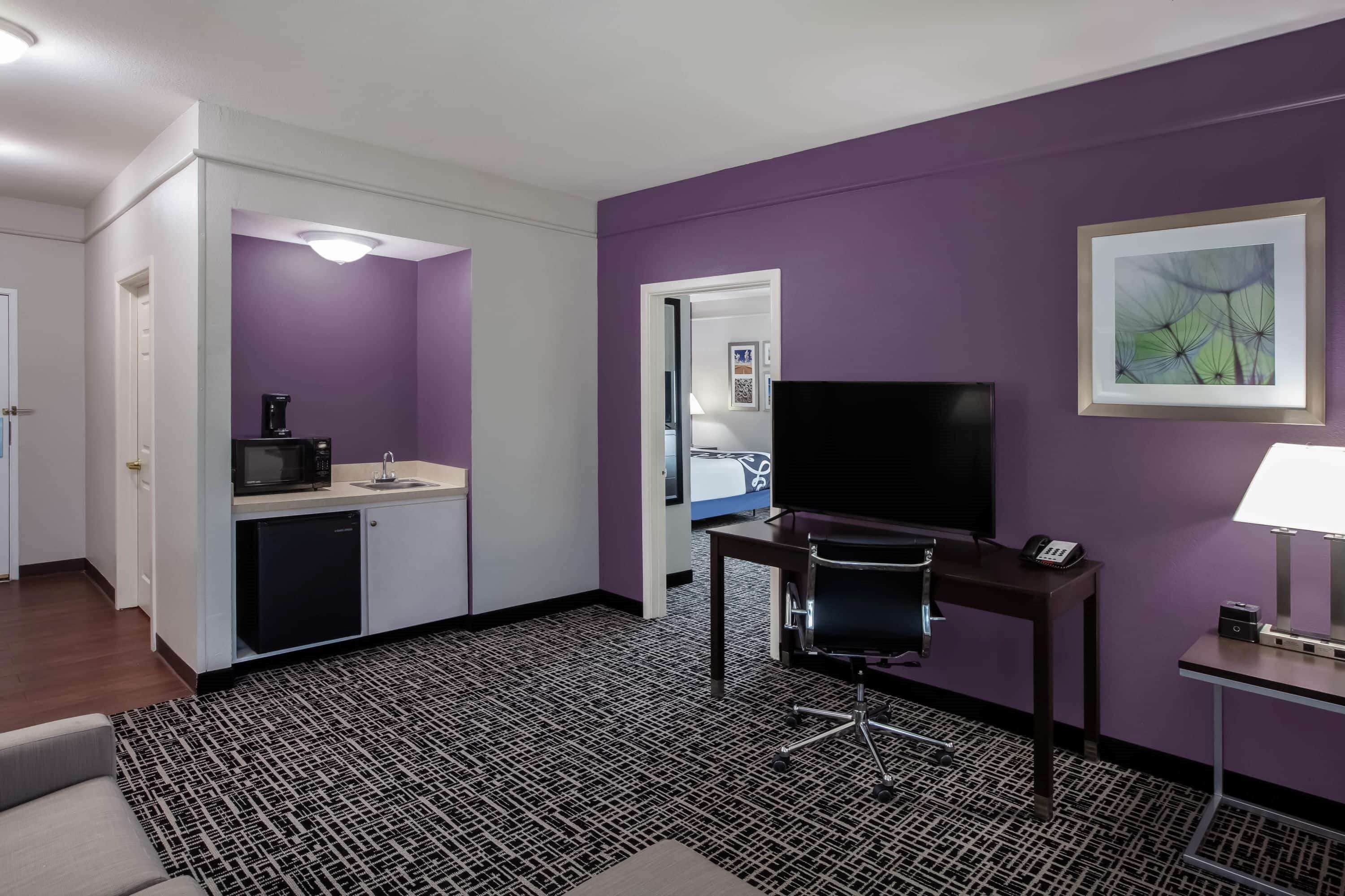 A hotel room with a kitchenette, a desk with a chair, a TV, and a framed picture on the wall.
