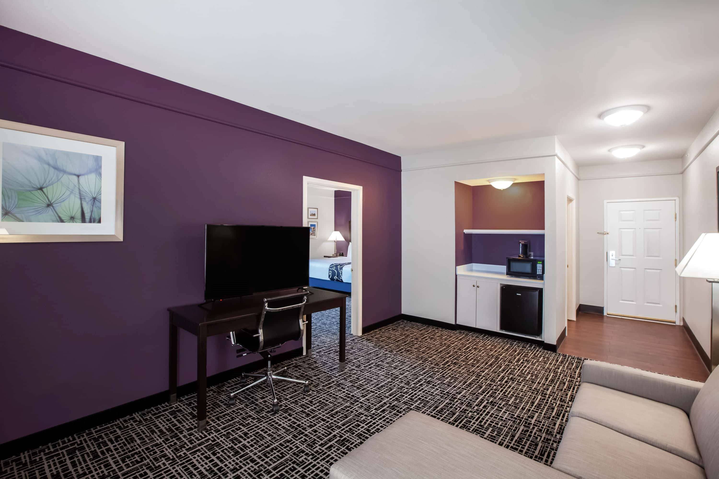 Une chambre d’hôtel avec un mur violet, une télévision, un bureau avec une chaise, un canapé, un coin cuisine avec un four à micro-ondes et une porte.
