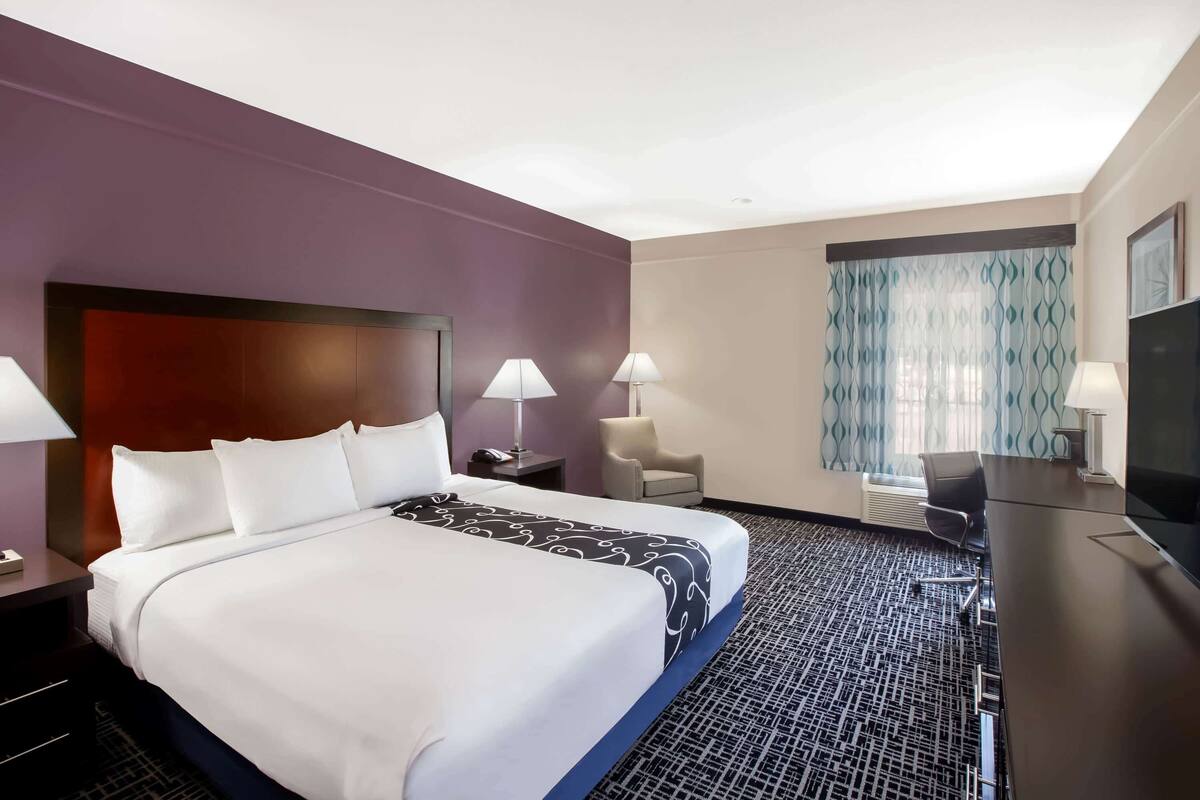 A hotel room with a large bed, two bedside lamps, a desk with a chair, a television, and a window with patterned curtains.