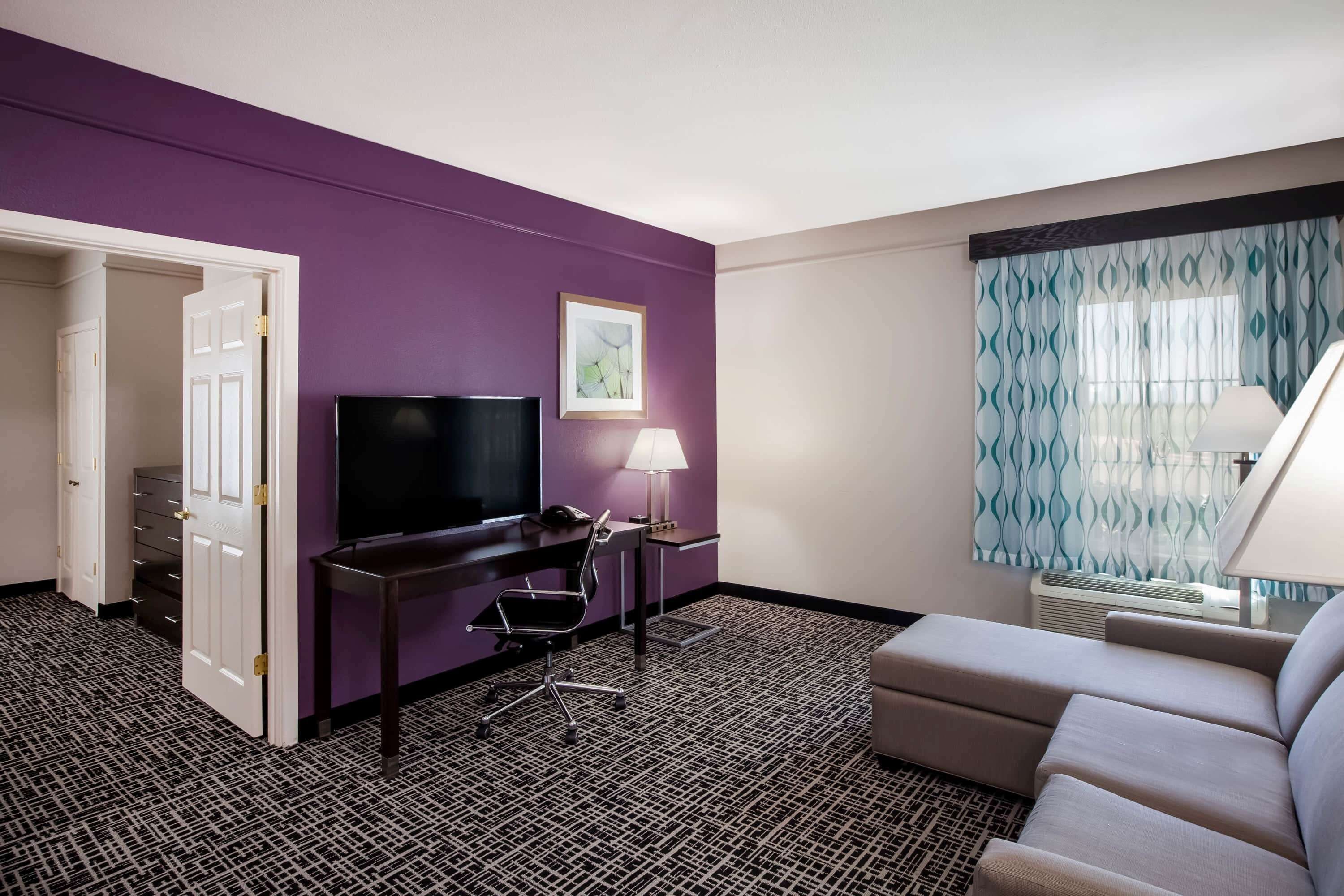 Une chambre d’hôtel avec un mur violet, un téléviseur à écran plat, un bureau avec une lampe, un canapé et une moquette à motifs.