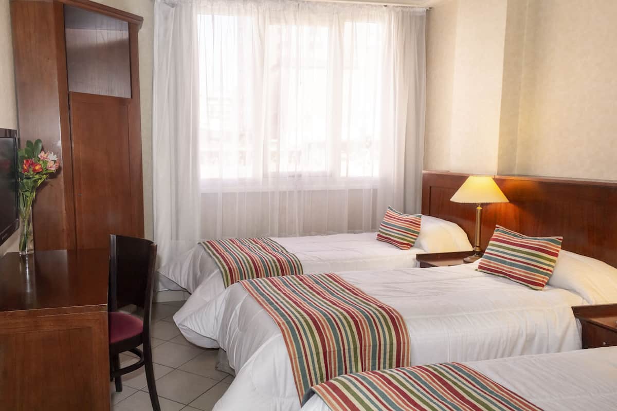 Habitación de hotel con tres camas, un escritorio, una silla, una lámpara y una ventana con cortinas traslúcidas.