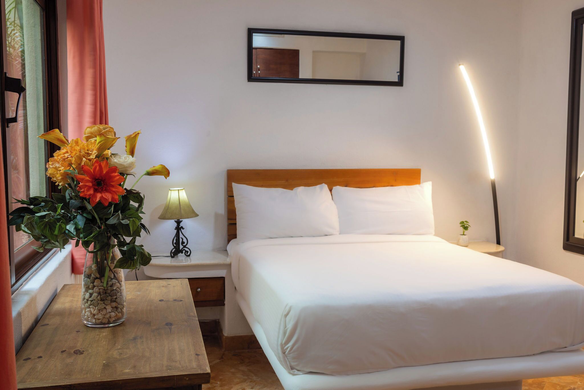 A hotel room with a bed, a nightstand, a lamp, a vase with flowers, and a mirror on the wall.