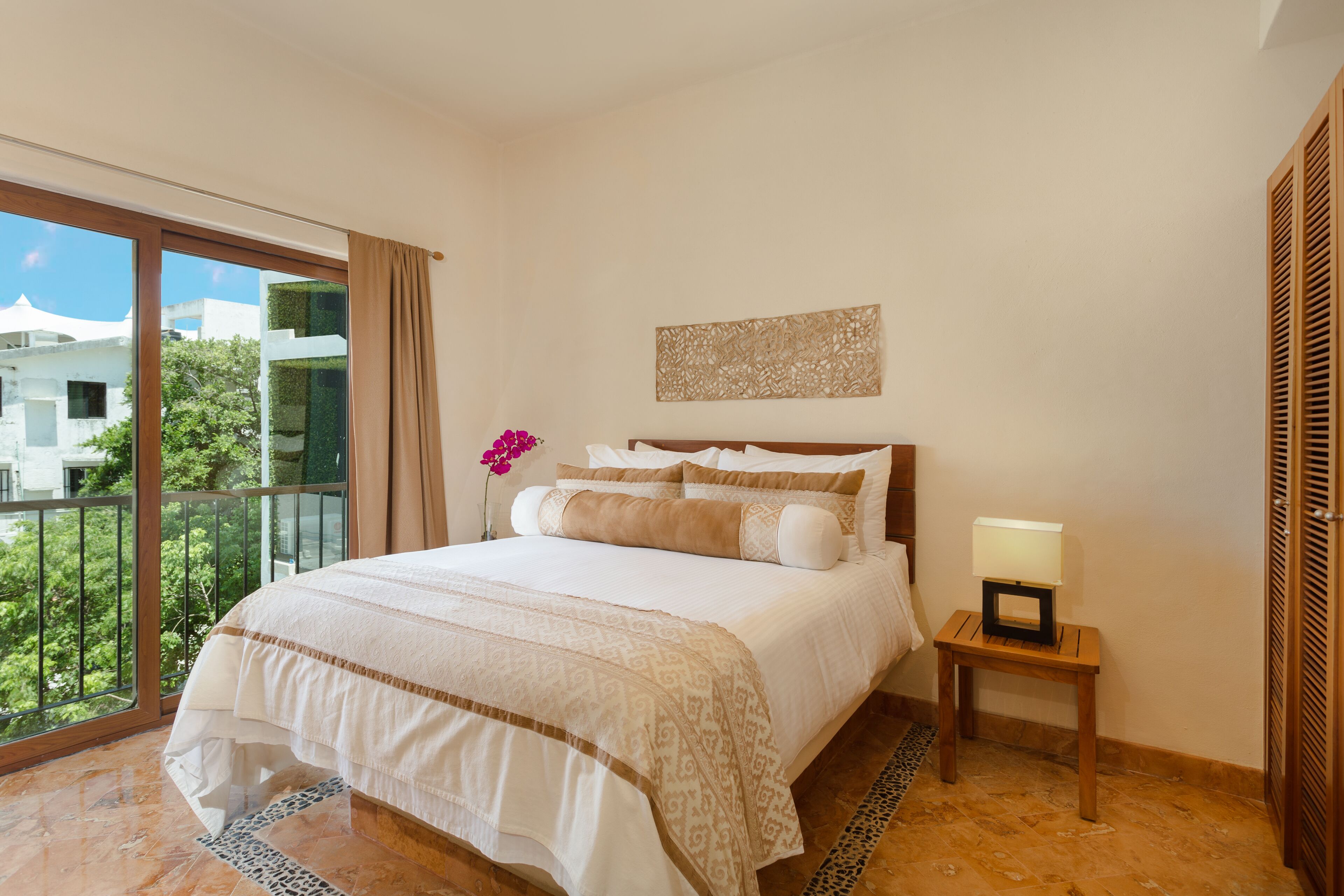 A hotel room with a large bed, a nightstand with a lamp, a view of greenery outside, and a decorative wall piece.