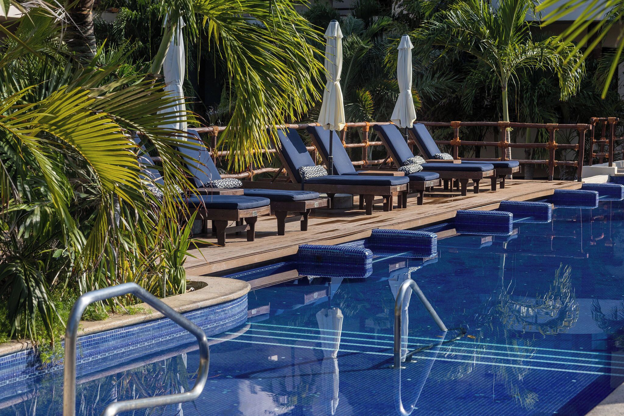 Un área junto a la piscina con sillones de descanso y sombrillas, rodeada de palmeras.