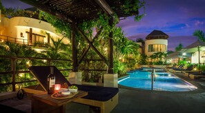Zona junto a la piscina con sillones, una mesa con una botella y aperitivos, y vistas a la piscina y los edificios del complejo.