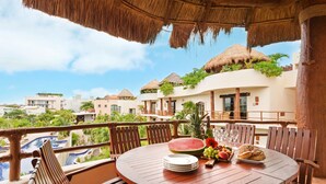 A round wooden table with plates, a watermelon, and a fruit tray on a balcony overlooking a swimming pool and resort buildings. - Porto Playa Condo Hotel & Beachclub (Playa del Carmen)