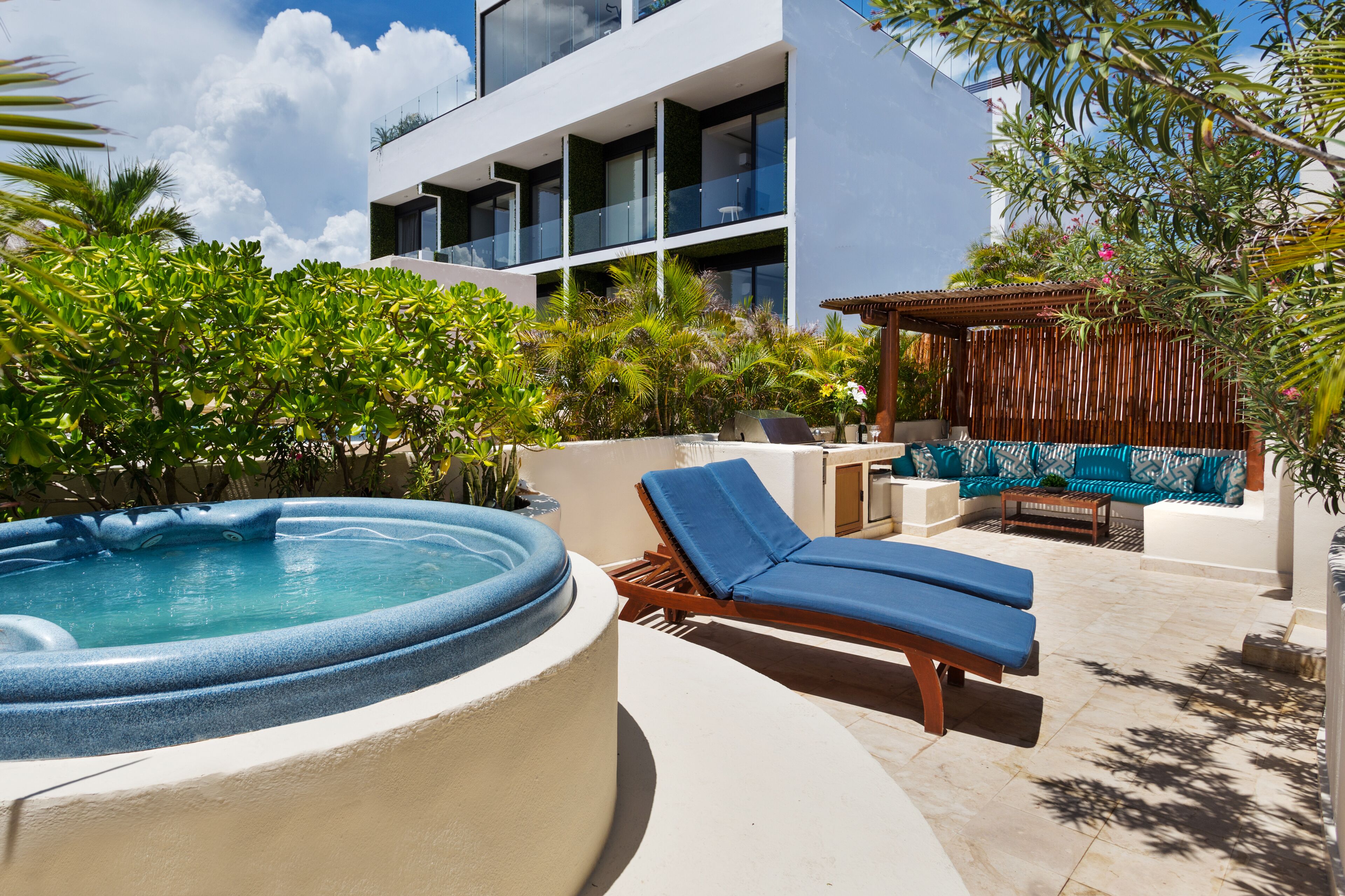 A modern outdoor pool area with lounge chairs and a hot tub, surrounded by lush greenery and a contemporary building in the background.