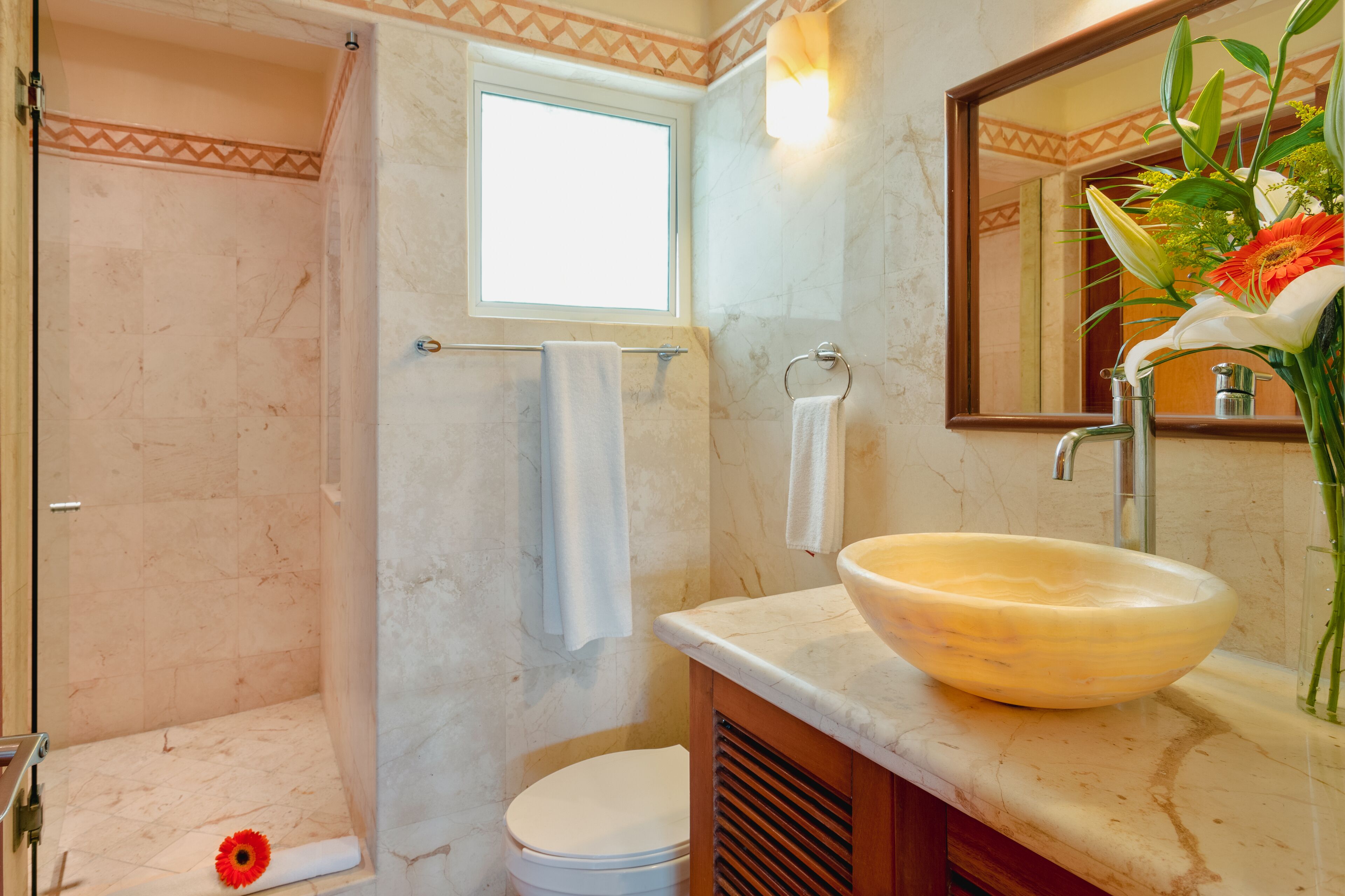 Un baño con piso de mármol, inodoro blanco, ducha con toallero, lavabo con recipiente grande y espejo con florero.