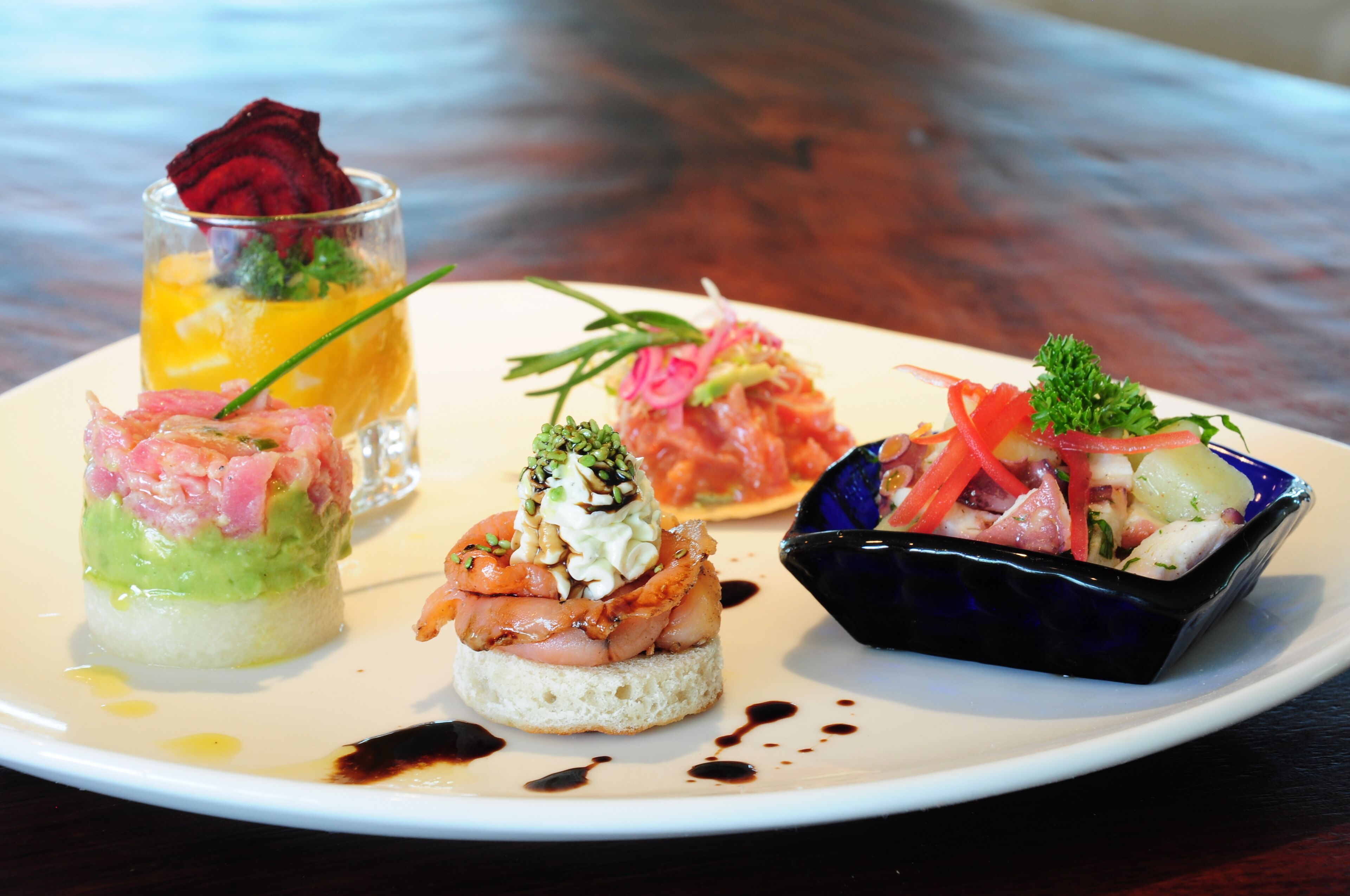A variety of appetizers on a white plate, including a glass with a red garnish, a dish with pink and green toppings, and a blue bowl with a salad.