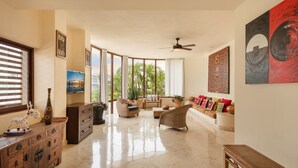 A spacious living room with a large window, a ceiling fan, and a variety of seating options. - Porto Playa Condo Hotel & Beachclub (Playa del Carmen)