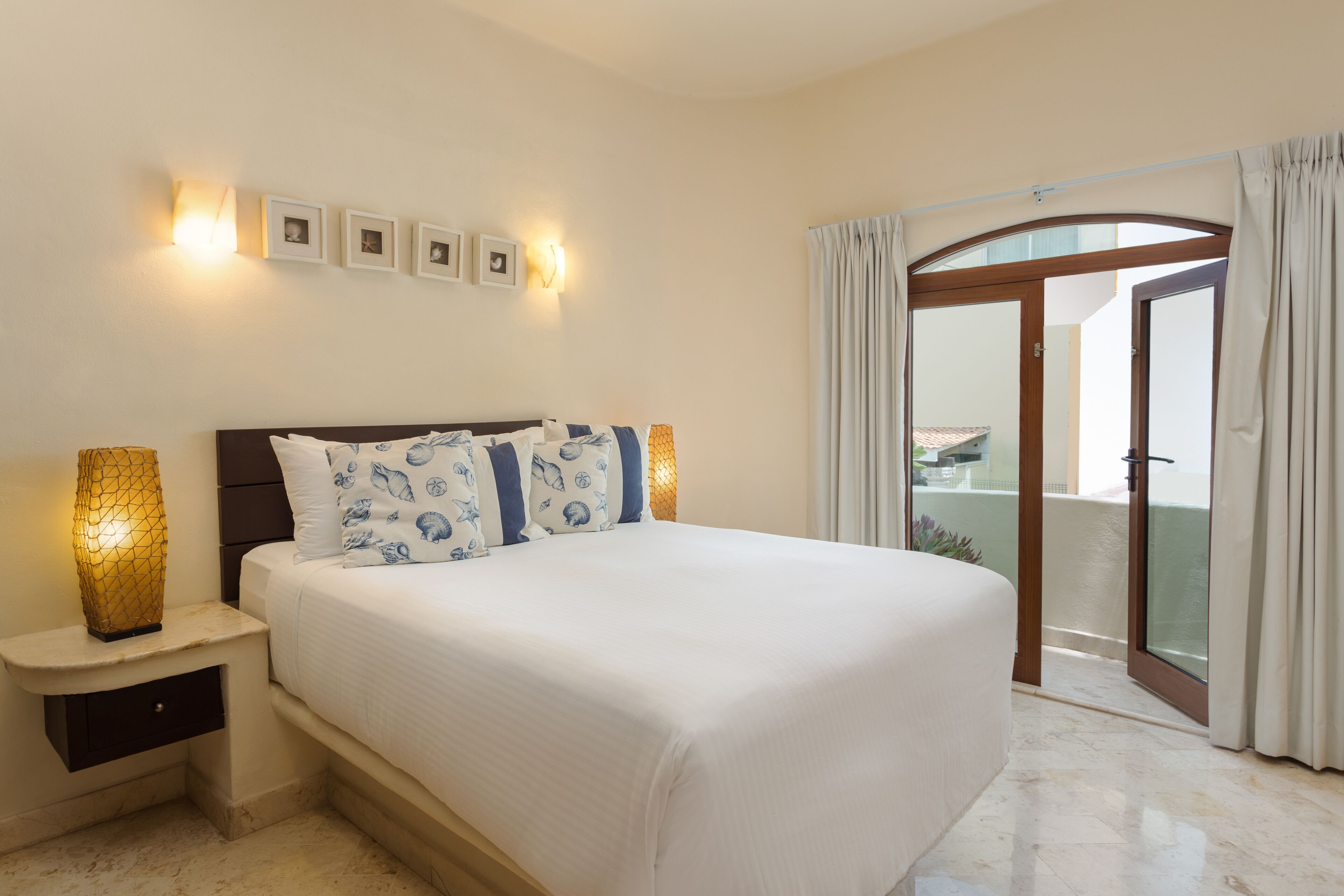 A bedroom with a bed, nightstands, wall-mounted lights, and a view through a glass door.
