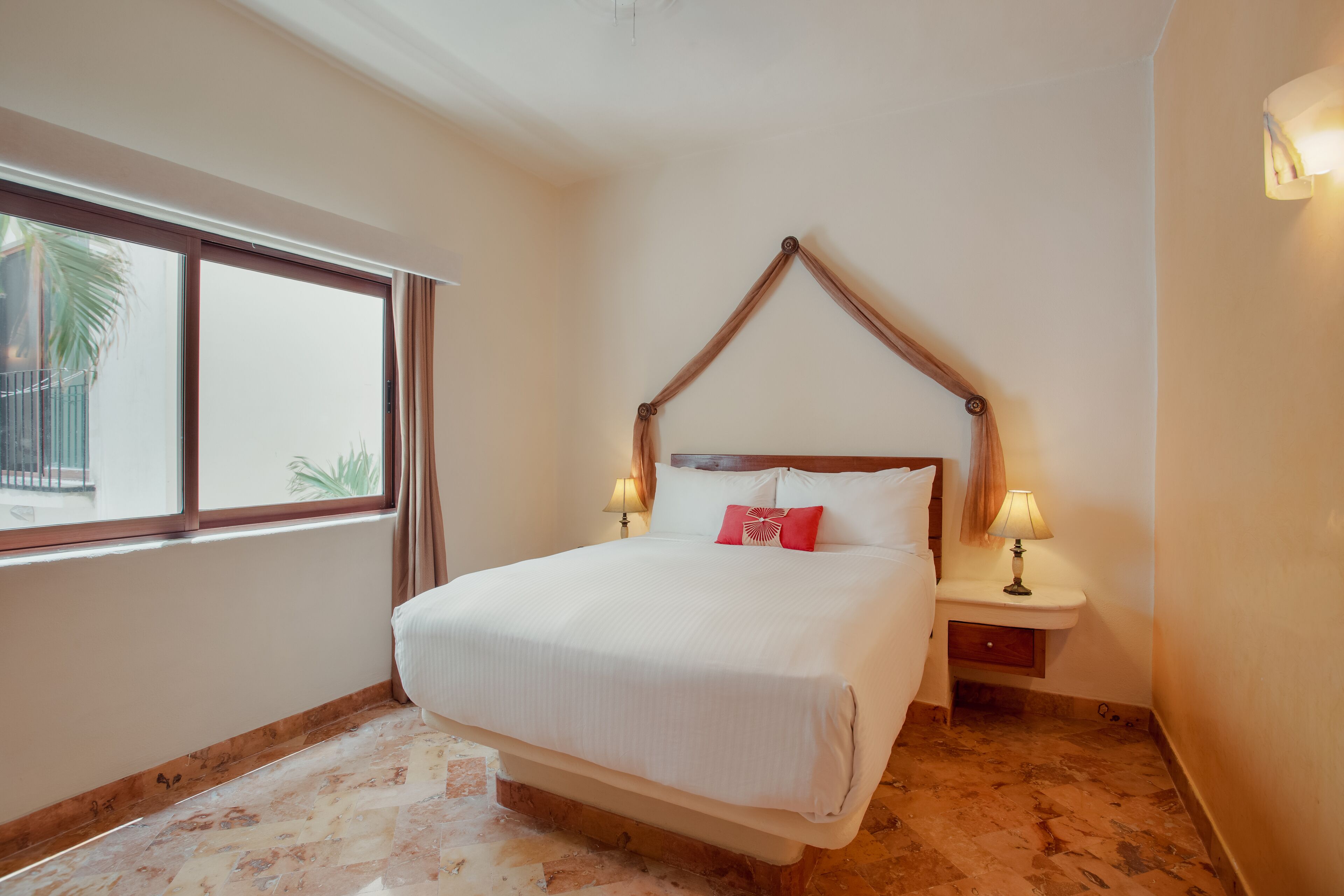 Una cama bien hecha con cabecera de madera, dos lámparas de noche y una ventana con cortinas.