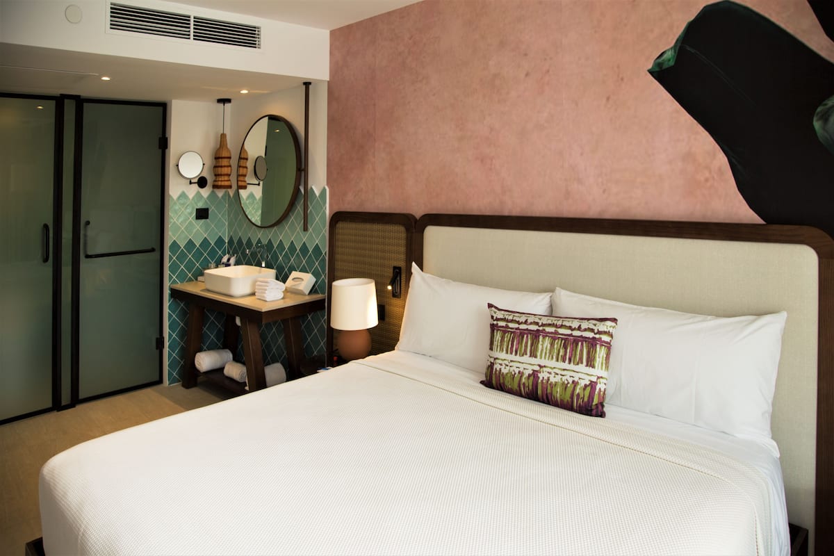 Habitación de hotel con una cama grande, una mesita de noche con lámpara, un espejo y un baño pequeño con lavamanos y toallero.